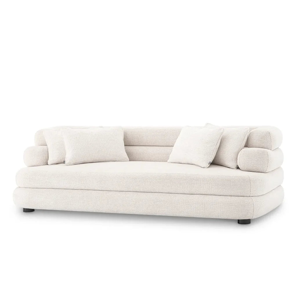 A modern low-profile sectional sofa upholstered in a light cream fabric, featuring four matching throw pillows and dark metal legs, shown against a plain white background.
