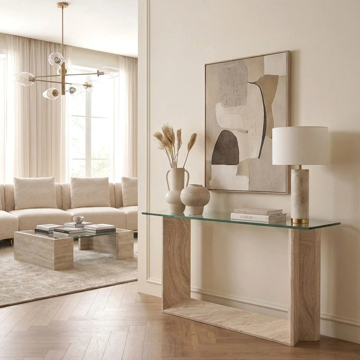 Console Table Fortuna. A modern glass console table with light wood legs in a bright, contemporary living room. The table is styled with a white table lamp, a vase with dried grasses, and a small ceramic pot, with a large abstract framed print hanging on the beige wall above it.