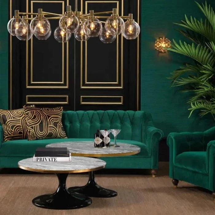Coffee Table Parme Round. A luxury living room setup featuring a tufted emerald green velvet sofa, a matching armchair, round marble-top coffee tables with gold trim, a modern multi-light brass chandelier, and a dark green wall with gold paneling.